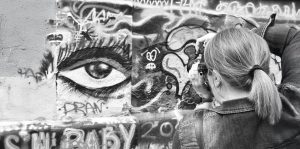 Schwarzweißfoto einer von hinten aufgenommenen Person. Die Person hat blonde, zu einem kurzen Zopf gebundene Haare, trägt eine Jeansjacke, ringförmige Ohrringe und eine Uhr am linken Handgelenk. Sie hält eine Kamera mit beiden Händen auf Augenhöhe und fotografiert die Wand vor sich, auf der das Graffiti eines Auges zu sehen ist.